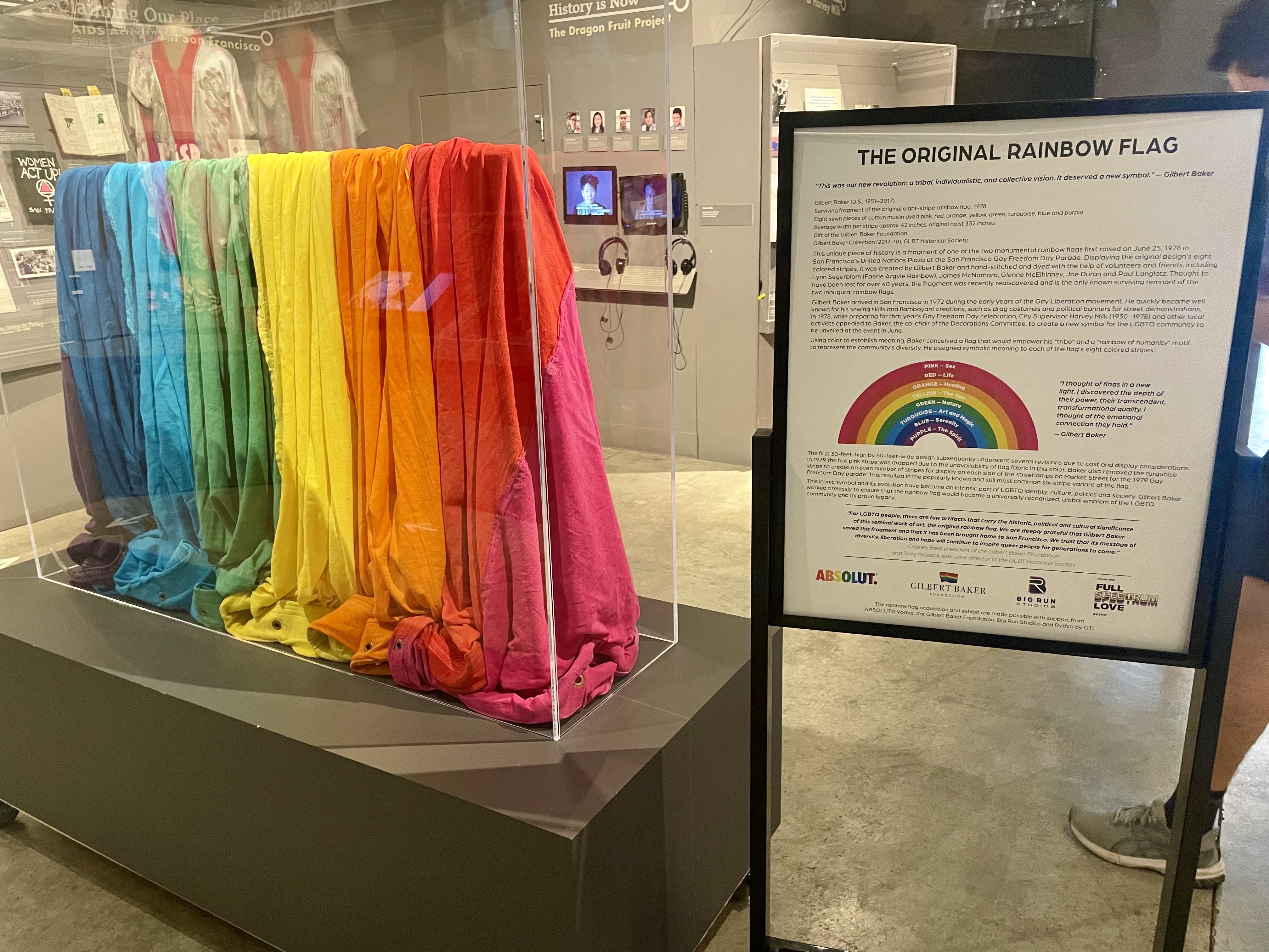 A museum display of the original rainbow pride flag, featuring eight vertical fabric panels in pink, red, orange, yellow, green, turquoise, indigo, and violet, housed in a clear protective case. To the right, a sign titled “The Original Rainbow Flag” explains the flag’s history, designer Gilbert Baker, and the meaning of each color. The exhibit is located in the GLBT Historical Society Museum in San Francisco.