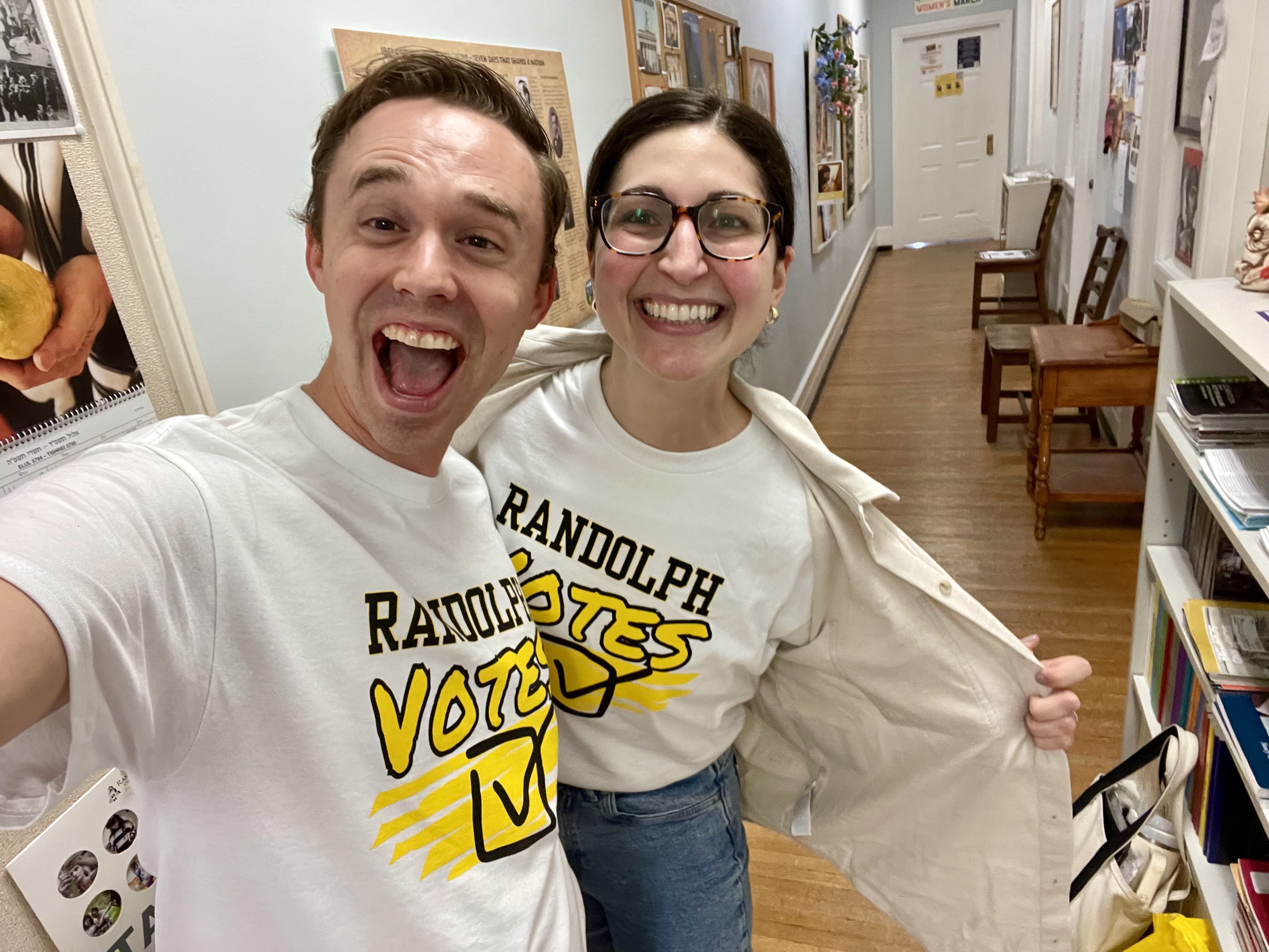Two people stand smiling widely in a brightly lit hallway, both wearing matching white T-shirts that say “Randolph Votes” in bold black and yellow lettering. One person points to their shirt with their coat open to reveal the design. The hallway is decorated with framed photos and posters, and the atmosphere is cheerful and energetic.