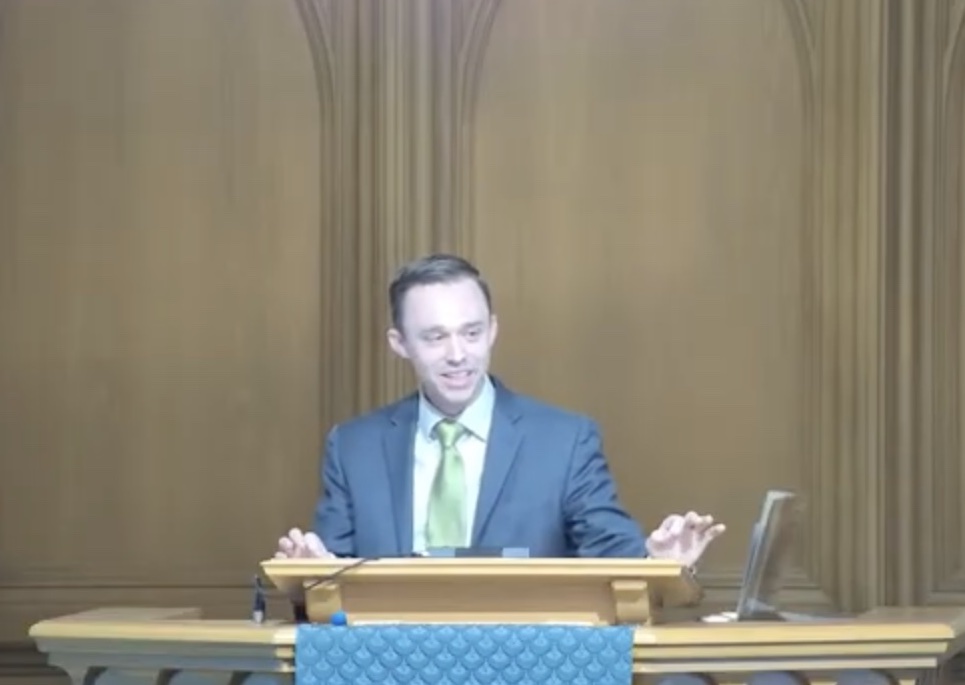 A photograph of Connor Kenaston delivering a sermon. He's standing in a pulpit wearing a green tie. Wood paneling is behind him.