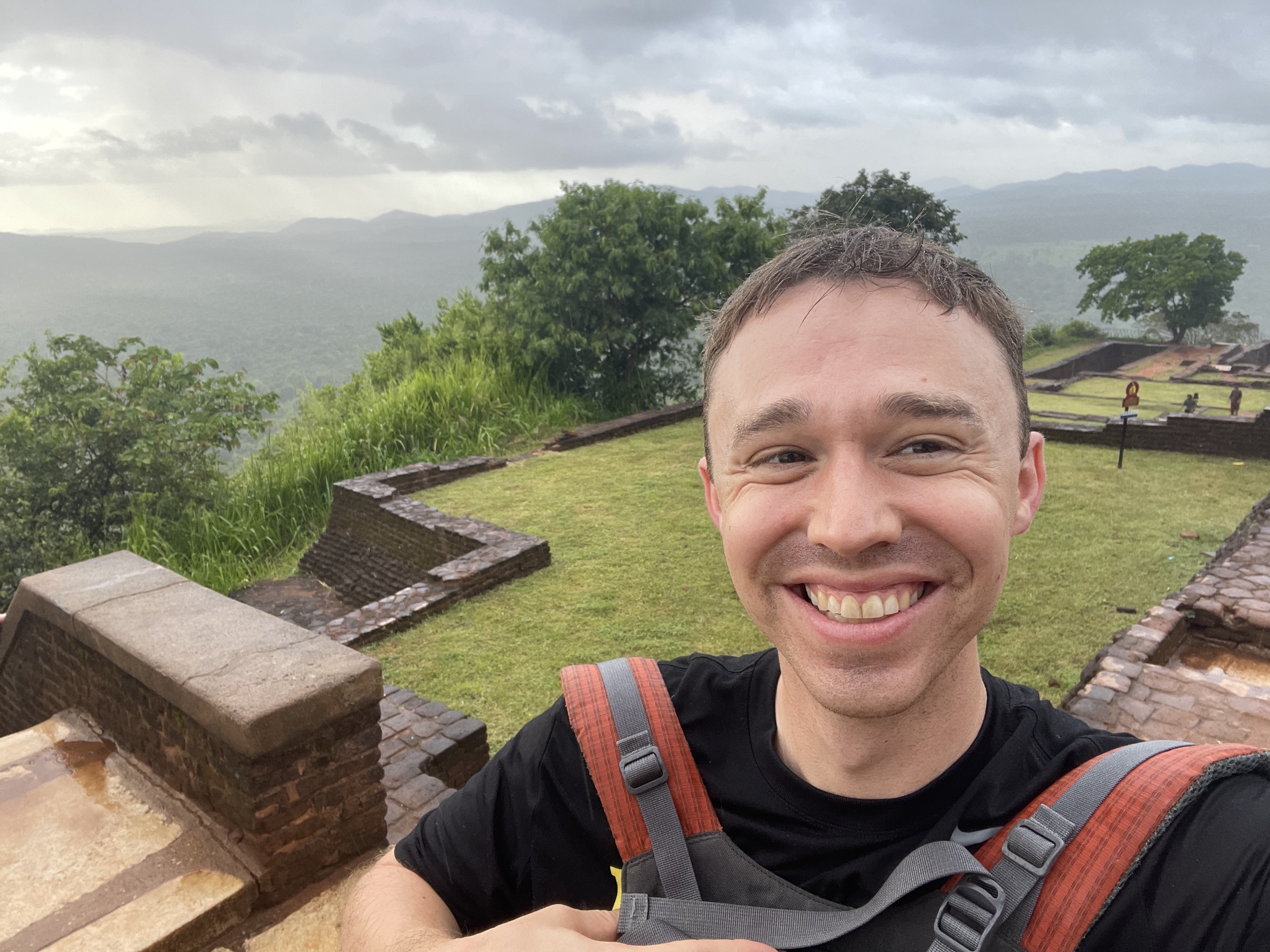 White man smiles toward camera. He's wearing an orange backpack and is standing on the top of Sigiriya with trees, mountains, and clouds in the background.